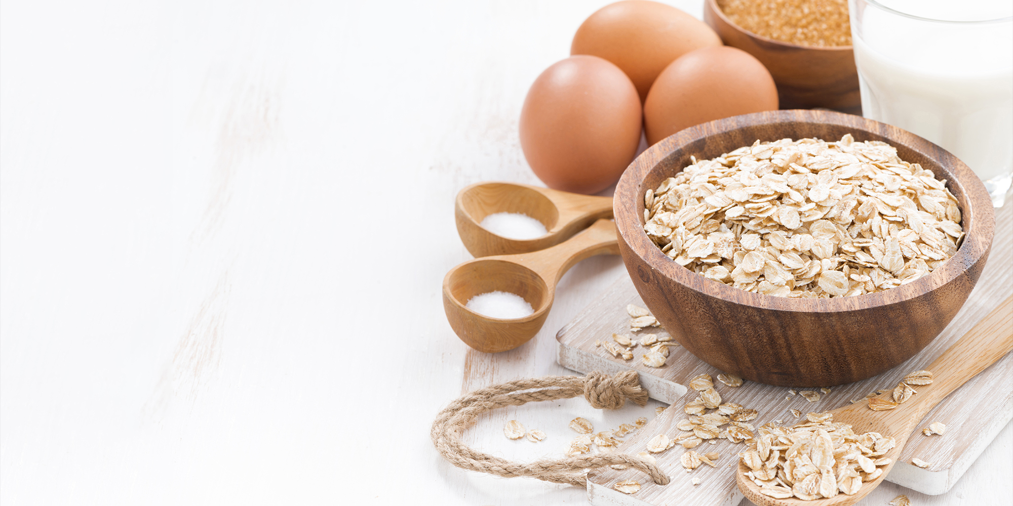 Wooden bowl filled with oats on chopping board. Various wooden spoons filled with oats or sugar. Eggs and glass of milk.